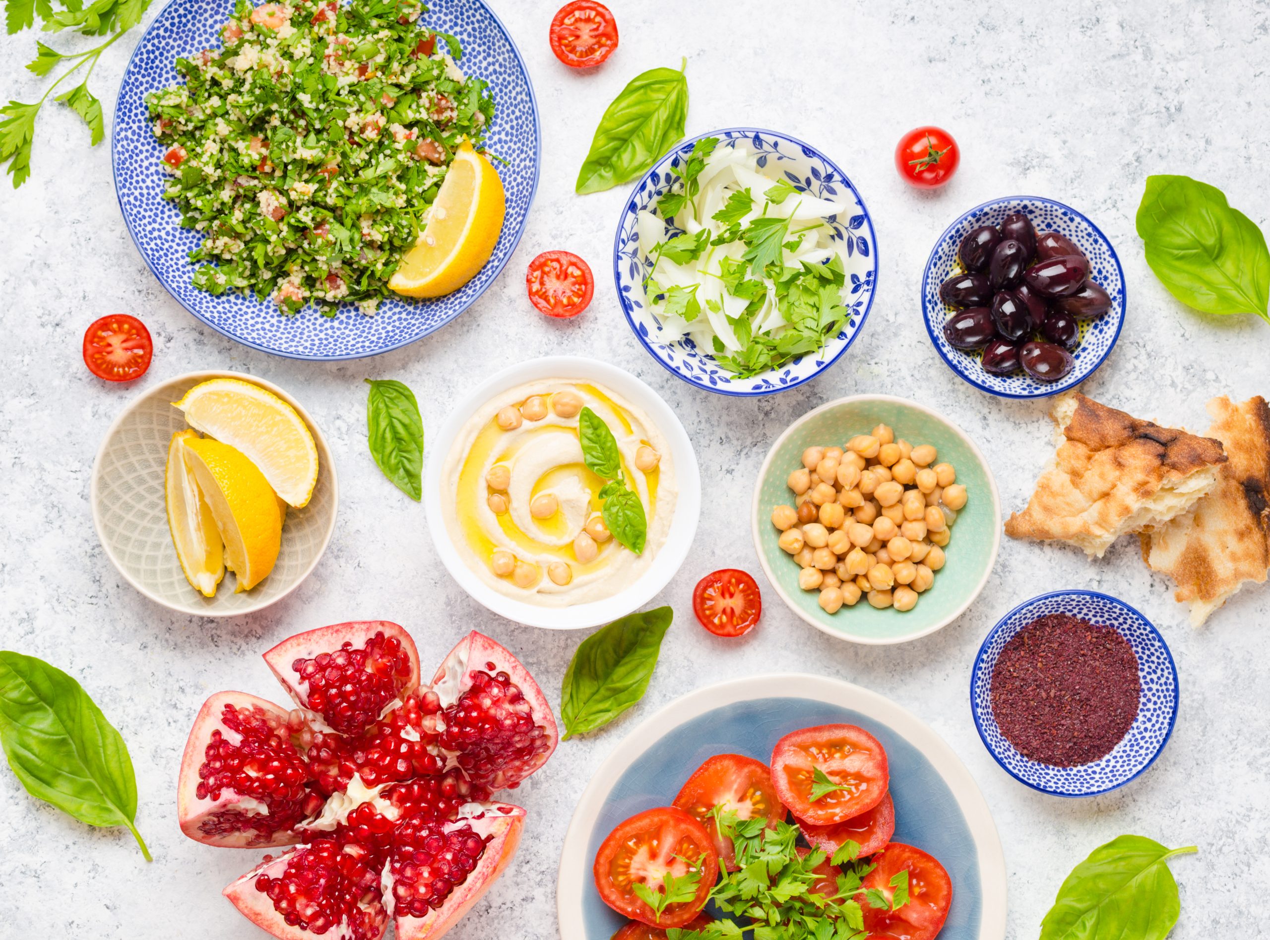 Top view of Middle eastern or arab dishes and assorted meze and snacks on concrete white rustic background: hummus, pita, tabbouleh vegetable salad, olives, chickpeas, tomatoes, pomegranate, herbs